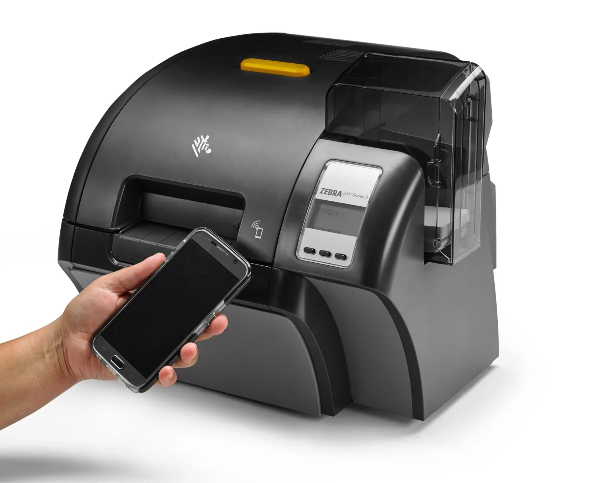 A Zebra T1200 thermal label printer in matte black with a small yellow top accent, shown on a white background. A human hand is holding a smartphone near the printer's display window, demonstrating a wireless connection feature. - Buy Zebra ZXP Printer With Lamination - Z93-000C0000EM00 in India