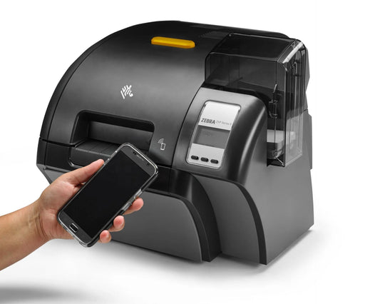 A Zebra T1200 thermal label printer in matte black with a small yellow top accent, shown on a white background. A human hand is holding a smartphone near the printer's display window, demonstrating a wireless connection feature. - Buy Zebra ZXP Printer With Lamination - Z93-000C0000EM00 in India